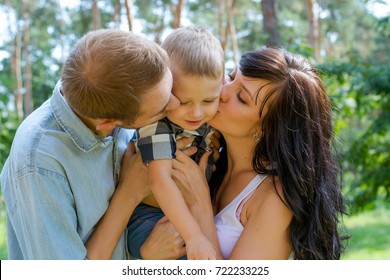 Mom And Dad Hug And Kiss Their Baby. Happy Family - Mom, Dad And Child- For A Walk In The Park. Family Resting In The Park In Sunny Day.