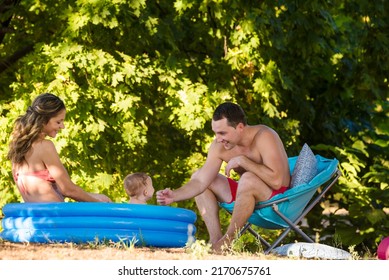 Mom, Dad And Little Boy Play In An Inflatable Pool Near A Sun Lounger On The Lawn. Family In Swimwear Tanning