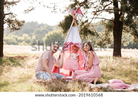 Mom, dad and little daughter are sitting next to wigwam decoration in the park. Family spending time outdoor in summer, having fun together. Girl are dressed in pink dress.