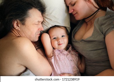 Mom And Dad Are Sleeping With The Child, Top View. The Baby Does Not Sleep And Peeps, And Tired Parents Sleep, The Baby Between The Parents. The Concept Of Parenthood And Breastfeeding