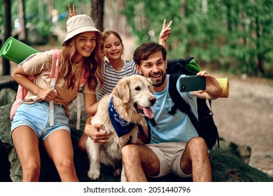 Mom and dad with their daughter with backpacks and a labrador dog take a selfie on the phone while sitting on a stone in the forest. Camping, travel, hiking. - Powered by Shutterstock