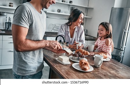 Mom, Dad And Their Little Beautiful Daughter Have Tea Party In The Kitchen And Talk. Father Puts Butter On Bread. Happy Family Concept.
