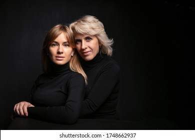 Mom And Daughter . Beautiful Woman 50 Years Old Blonde And Girl Hugging. They Are In Sweaters On A Black Background In The Studio. Emotional Family Photo.