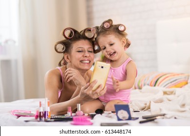 Mom And Daughter In The Bedroom On The Bed In The Curlers Make Up, Paint Their Nails And Have Fun