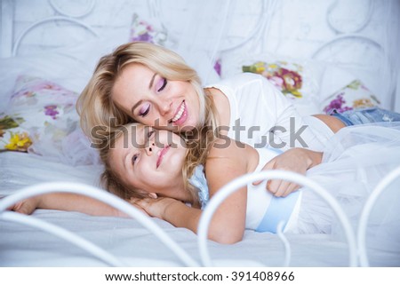 Mom and daughter blonde caucasians are happy and smiling on a white bed in a white room. Girl gently kisses her mom. Beautiful smile. Happy beautiful family. Mothers Day