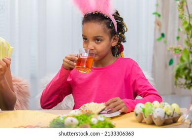 Mom and daughter with funny bunny ears are drinking tea with Easter cake. African American woman and little girl are sitting at table in festively decorated room at home. Happy easter. Close up.