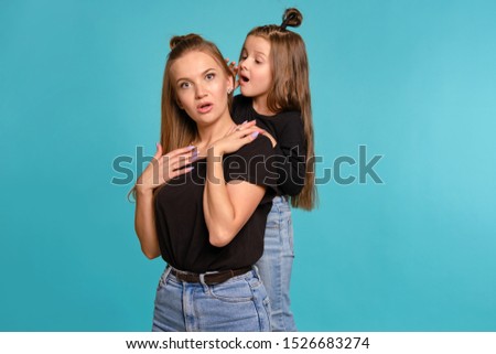 Mom and daughter with a funny hairstyles, dressed in black shirts and blue denim jeans are posing against a blue studio background. Close-up shot.