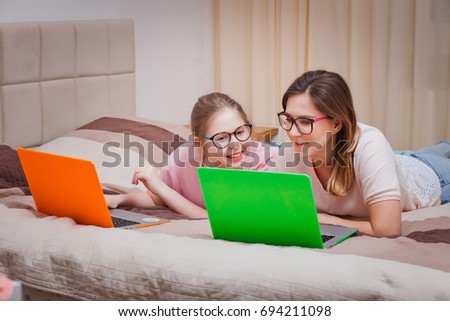 Mom and daughter in glasses lie on the bed in the bedroom and use two bright laptops and have fun