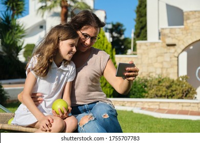 Mom And Daughter Kid Using Smartphone For Video Call. Family Sitting On An Outdoor Chair On Lawn In The Yard