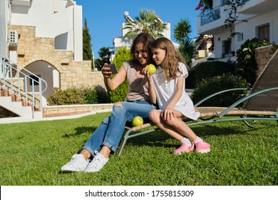 Mom And Daughter Kid Using Smartphone For Video Call. Family Sitting On An Outdoor Chair On Lawn In The Yard