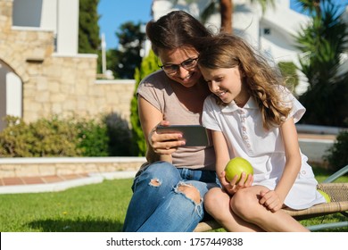 Mom And Daughter Kid Using Smartphone For Video Call. Family Sitting On An Outdoor Chair On Lawn In The Yard