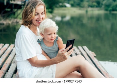 Mom And Daughter Look At The Phone And Laugh On A Pier By A Lake In The Countryside In Summer. Spending Time Together And Having Fun. Family Pleasure Activity