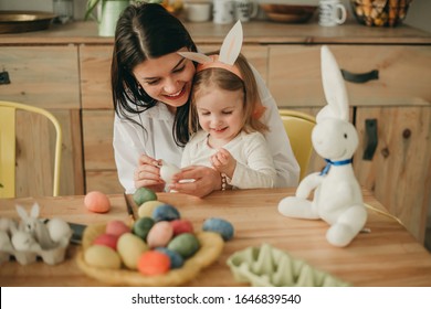 Mom And Daughter Paint Easter Eggs In The Kitchen With Tassels, Looking At The Phone, Tulips And Rabbits