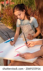 Mom And Daughter Paint A Picture Together Outdoors In The Park. A Little Girl In The Spotlight, Drawing Strokes With Acrylic Paint For The Future Picture. Photography In Warm Colors