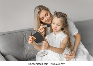 Mom and daughter play with cosmetics in a cozy home. A young woman and a girl in white linen dresses sit on the couch and make up for each other. - Powered by Shutterstock