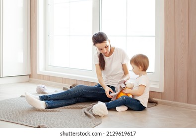 Mom And Daughter Play Dress Up Soft Toy At Home Next To Window In Bright Modern Interior. Mother And Small Child Spend Time Together, Lifestyle