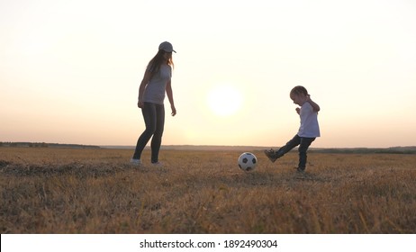 Mom And Daughter Play Football On Field In Rays Of The Sunset. Teamwork Of The Child And Mom. Happy Family And Child Have Fun Playing A Soccer Ball In Park. The Kid Kicks Ball. Healthy Sports Family