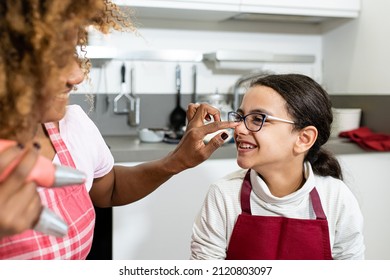 Mom And Daughter Play While Cooking, Latin American Mother Dirty With Pink Cream On Little Girl’s Nose, Funny And Playful Scene In Family