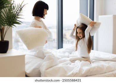 Mom and daughter playing together in bed, fighting with pillows, creating joyful memories. Happy family moment in bright modern bedroom with large windows, natural light, and indoor plant. - Powered by Shutterstock