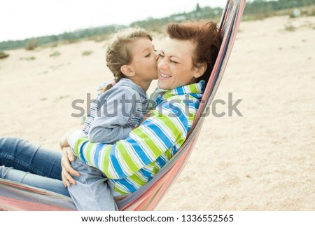 Mom and daughter sit in a hammock on the beach smiling and daughter kisses mom on the cheek