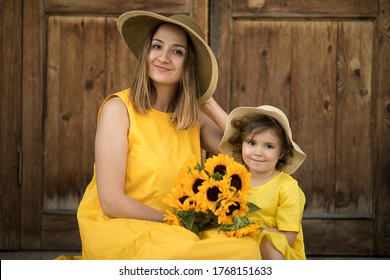 Mom With A Daughter Are Sitting On A Wooden Porch. Mom With A Daughter In Yellow Dresses And Straw Hats. Mom Holds A Bouquet Of Sunflowers. Image With Selective Focus.