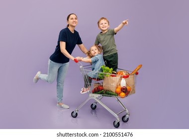 Mom And Daughter And Son Point To Something While Shopping, A Girl And A Boy Are Sitting In A Grocery Cart. Isolated Concept Image Of The Shopping With Cjpe Space.