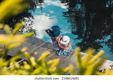 Mom And Daughter In Summer Dresses And Hats On Their Heads Enjoy The Pier With A Boat By The Water