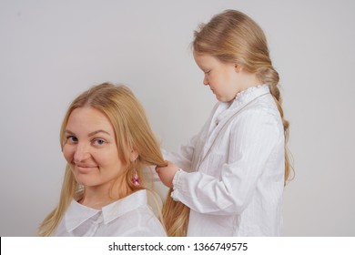 Mom And Daughter In White Shirts With Long Blonde Hair Posing On A Solid Background In The Studio. A Charming Family Takes Care Of Each Other And Makes Braids.