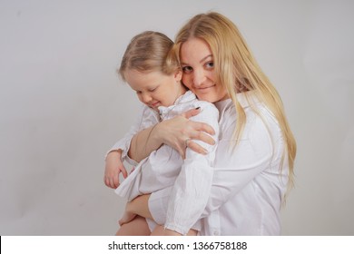 Mom And Daughter In White Shirts With Long Blonde Hair Posing On A Solid Background In The Studio. Charming Family Takes Care Of Each Other And Hugs.