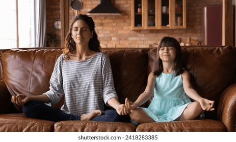 Mom and daughter yogis. Calm hispanic mother and little girl practice yoga exercise sit on sofa in lotus pose breath deep relax. Preteen child imitate elder sister nanny in doing meditative gymnastics - Powered by Shutterstock