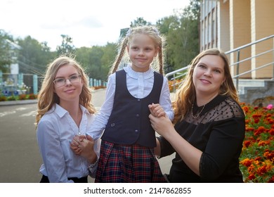 Mom And Daughters Who Are Schoolgirls In Uniform Near The School In Russia On September 1. Mother And Sisters Girls In City In Serious Clothes