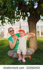 Mom Doing Yoga With Your Child. Baby Crawling On The Grass On A Background Of Sitting His Mom. The Kid Runs Away From His Mother.