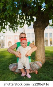 Mom Doing Yoga With Your Child. Baby Crawling On The Grass On A Background Of Sitting His Mom. The Kid Runs Away From His Mother.