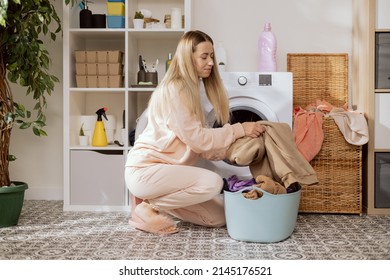 Mom dressed in sweatpants does housework in the bathroom, laundry room, sits on the floor with a blue large bowl filled with clothes at the washing machine, loads colorful things into the drum. - Powered by Shutterstock