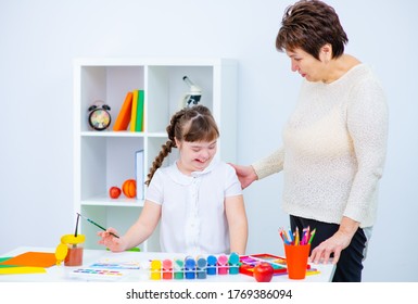 Mom Encourages A Child With Down Syndrome During Creative Activities. Teacher And Student With Disabilities In Class At A Special School