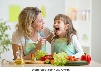 Mom Feeding Child Daughter Vegetables In Kitchen