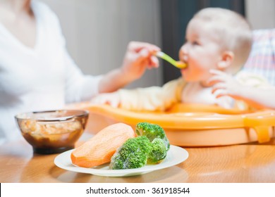 Mom Feeds The Baby Soup. Healthy And Natural Baby Food. Vegetables, Carrots, Cabbage, Broccoli. Child Sitting On The Highchair At The Table.