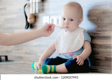 Mom Feeds Her Beautiful Baby With A Spoon, The Child Eats Porridge, Against The Background Of An Ordinary Apartment In The Frame You Can See The Hand Of Mom And Baby Who Eats