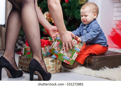 Mom Gives A Bad Gift. The Child Sits On A Box With Gifts At The Christmas Tree And Fireplace.