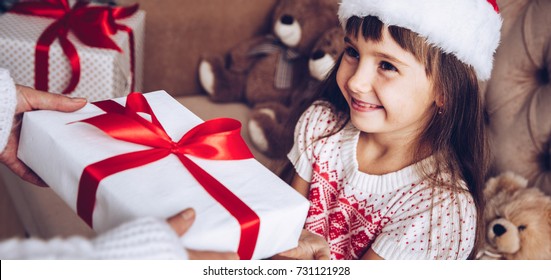 Mom Gives A Gift To Her Daughter, Christmas Kids,child, Portrait.Little Girl In Santa Claus Hat At Home With Xmas Tree.Close Up,selective Focus.Receive A Present.