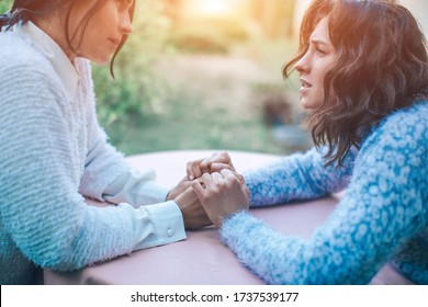 Mom Gives Her Daughter Advice. Sunny Glare. Two Young Beautiful Brunette Women Are Sitting At A Round Table Holding Hands And Holding Each Other. A Very Important Feeling Of Feminine Support Is Care. 