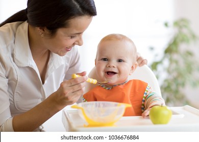 Mom giving homogenized food to her baby son on high chair in kitchen.