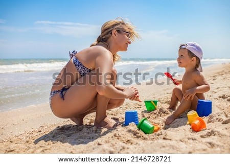 Mom with glasses plays with a little toddler sitting on the beach near the sea