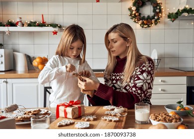 Mom Helping Cute Small Kid Daughter Making Christmas Cookies Preparing Gifts Together Sitting At Kitchen Table. Mother And Child Girl Making Family Xmas Holiday Gingerbread Decorations At Home.