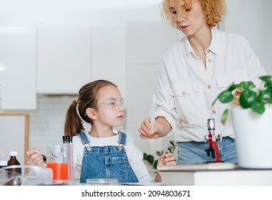 Mom Helping Little Girl To Do A Home Science Project, Handing Her A Flask. She Has Chemistry Glassware With Colorful Liquids And Microscope