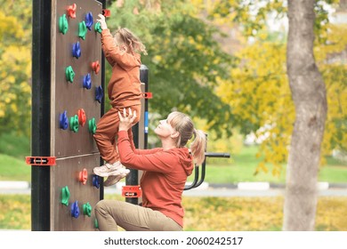 Mom Helps Her Little Daughter To Overcome The Artificial Mountain Relief On The Street Wall For The Climbing Of The Sports Ground. Sport And Family Leisure Concept