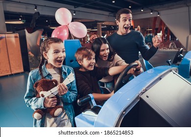 Mom Helps Her Son Drive A Car In A Computer Game. Dad And Daughter Support Mom And Son Driving A Toy Car In A Video Game. 