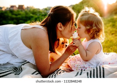 Mom With Her 2 Years Old Daughter Sitting On The Blanket And Eating Ice Cream In The Park. Sunset. Cute Dog Looking On Them. Good Relations Of Parent And Child. Happy Moments Together.