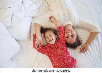 Mom With Her 6 Years Old Little Daughter Dressed In Winter Pajamas Are Relaxing And Playing In The Bed At The Weekend Together, Lazy Morning, Warm And Cozy Scene. Top View Point.