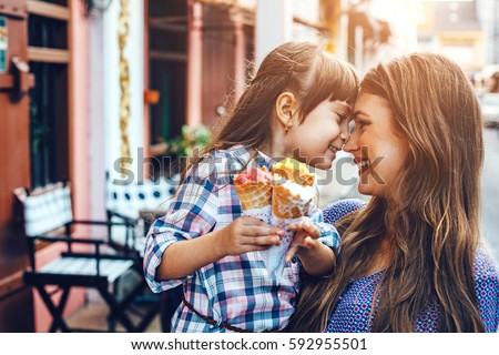 Mom with her 6 years old daughter walking along city street and eating ice cream in front of the outdoor cafe. Good relations of parent and child. Happy moments together.
